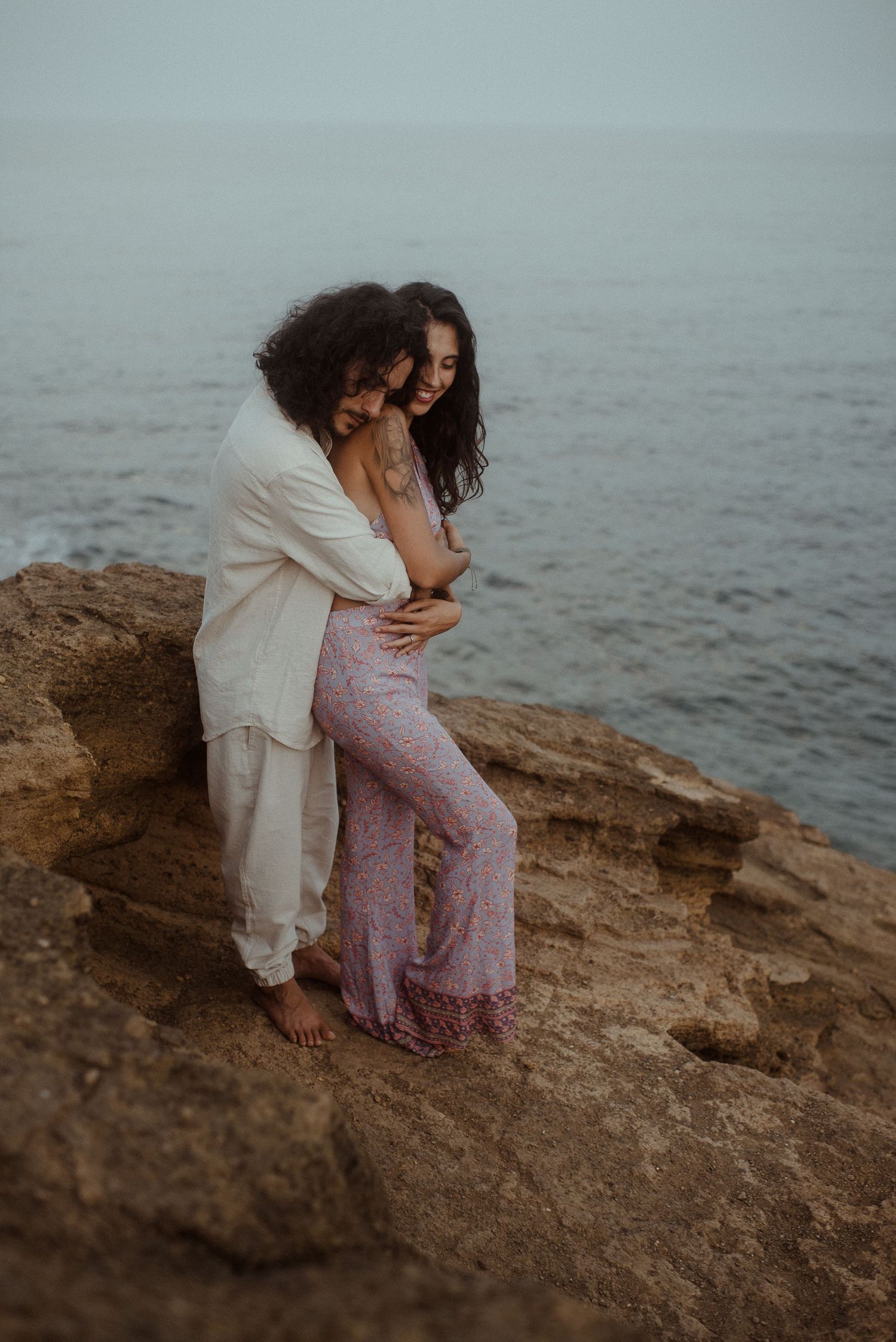 A loving couple holds each other close while standing on rocky ocean cliffs.