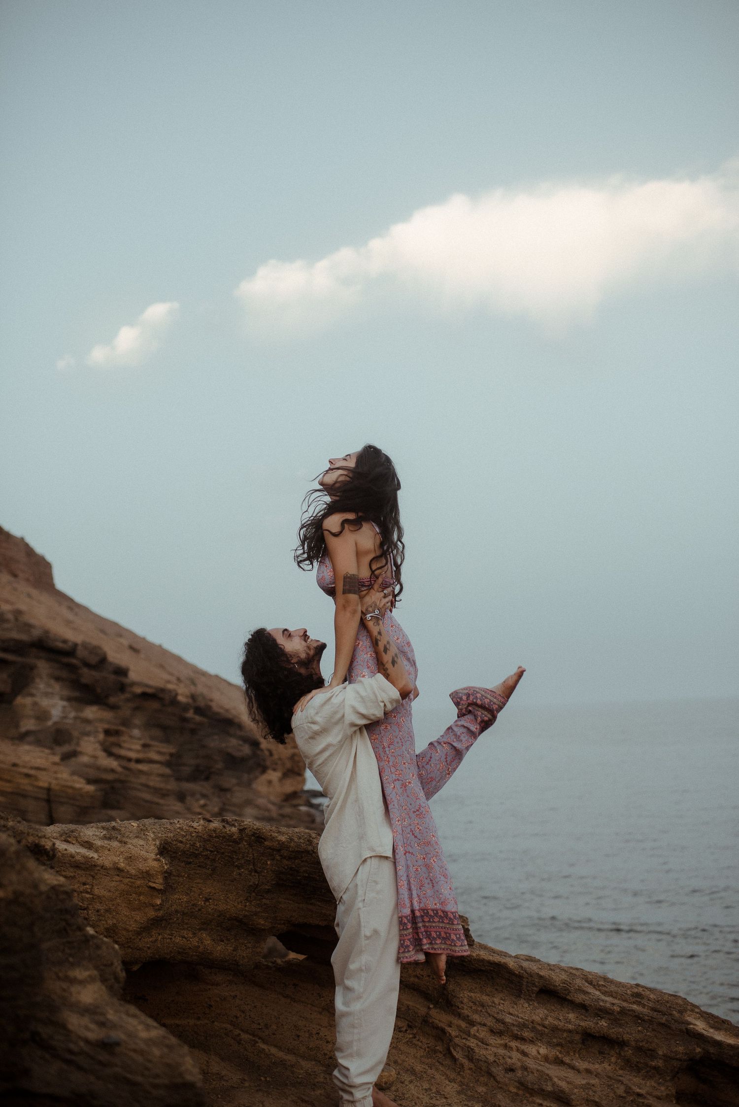 A couple shares a joyful moment as one lifts the other on coastal cliffs by the ocean.