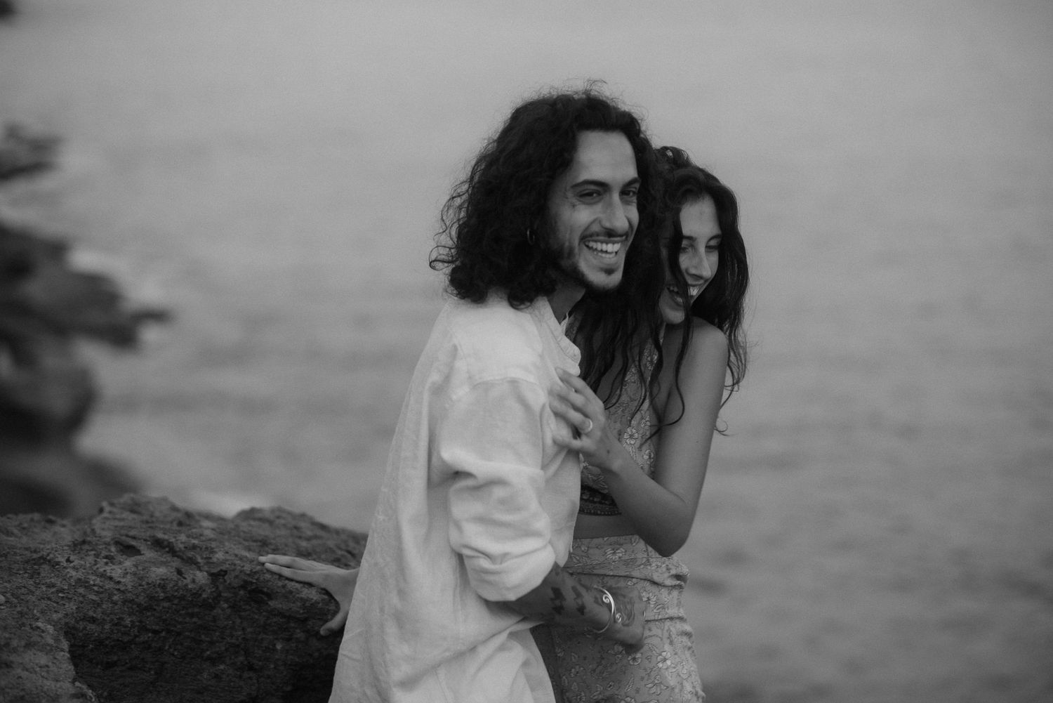 A couple laughs and embraces while standing on coastal rocks with the ocean in the background.