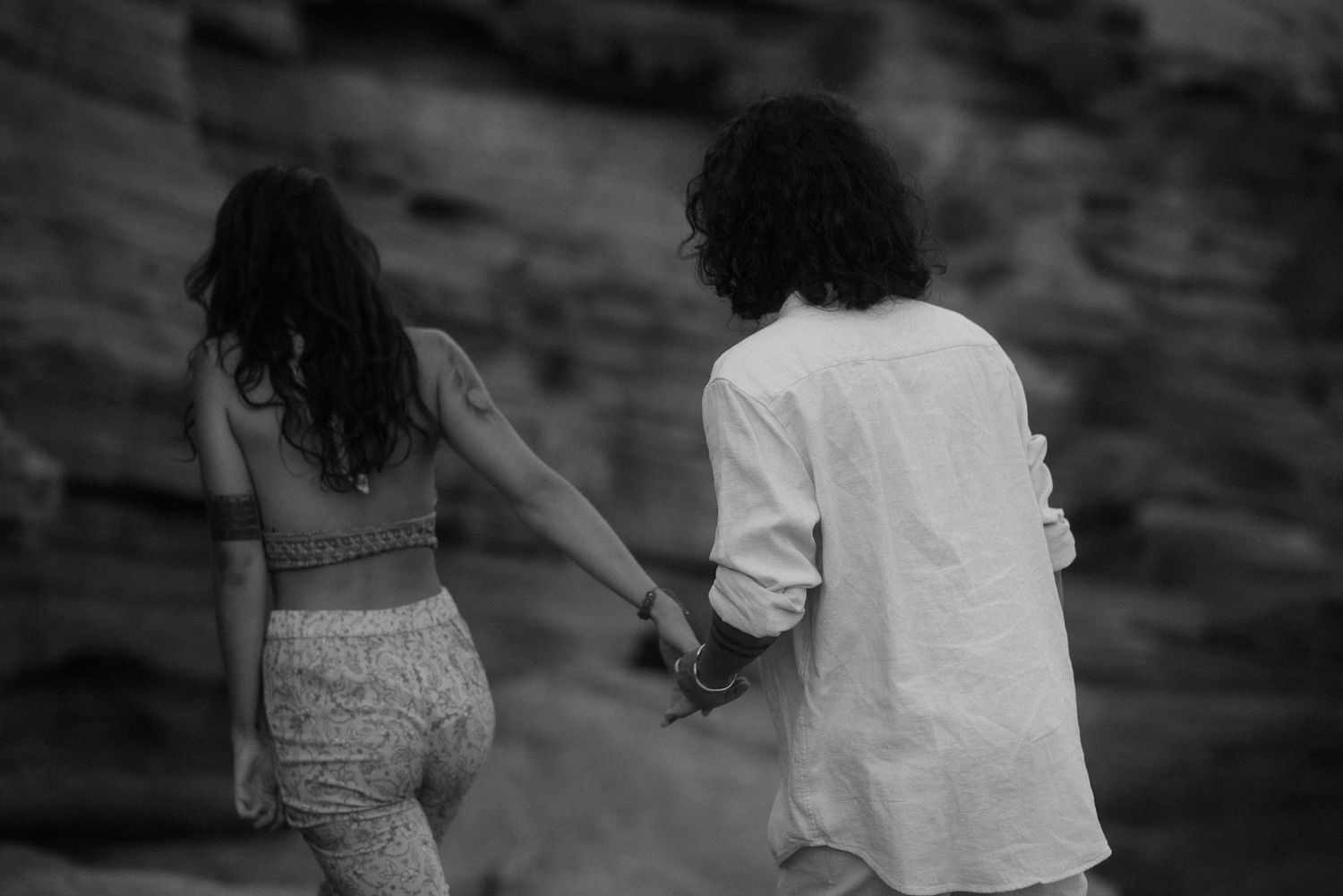 A couple holding hands walks along dramatic coastal cliffs in black and white.