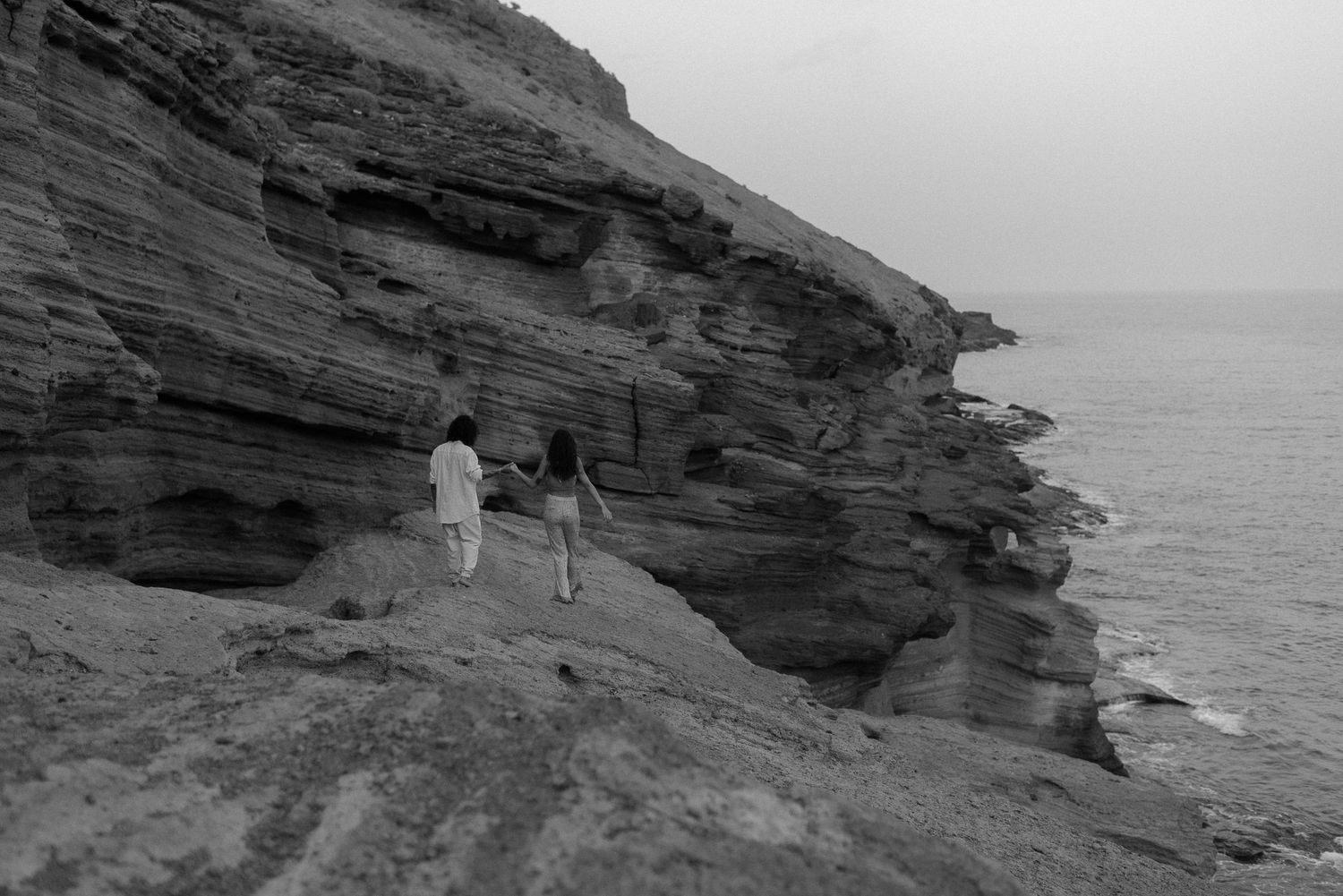 Two figures stand on dramatic rocky cliffs overlooking the ocean in black and white.