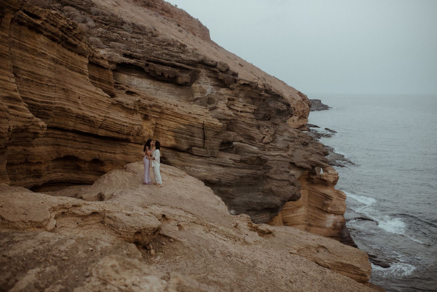 A couple stands together on rugged coastal cliffs during an overcast day.