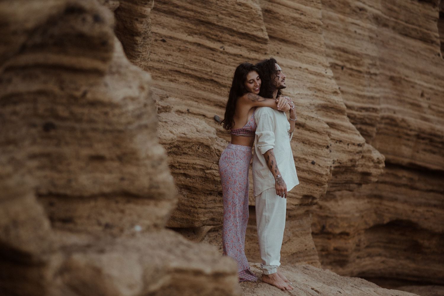 A couple shares an intimate moment against dramatic red rock cliffs by the ocean.