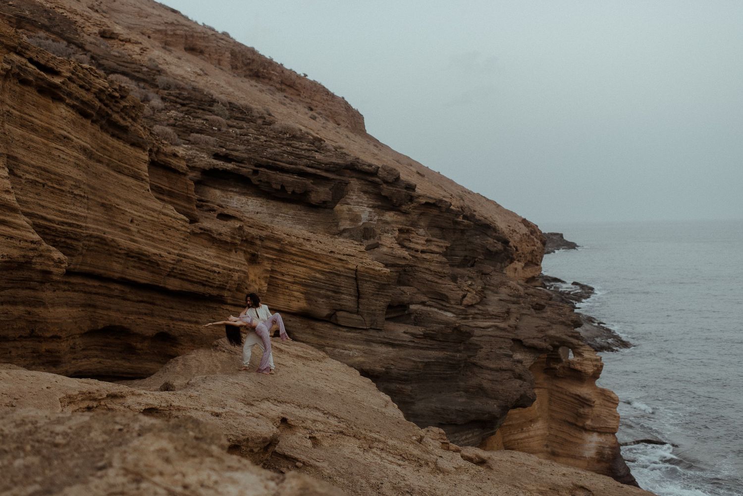 Couple embraces on dramatic rocky coastal cliff overlooking misty ocean at sunset.