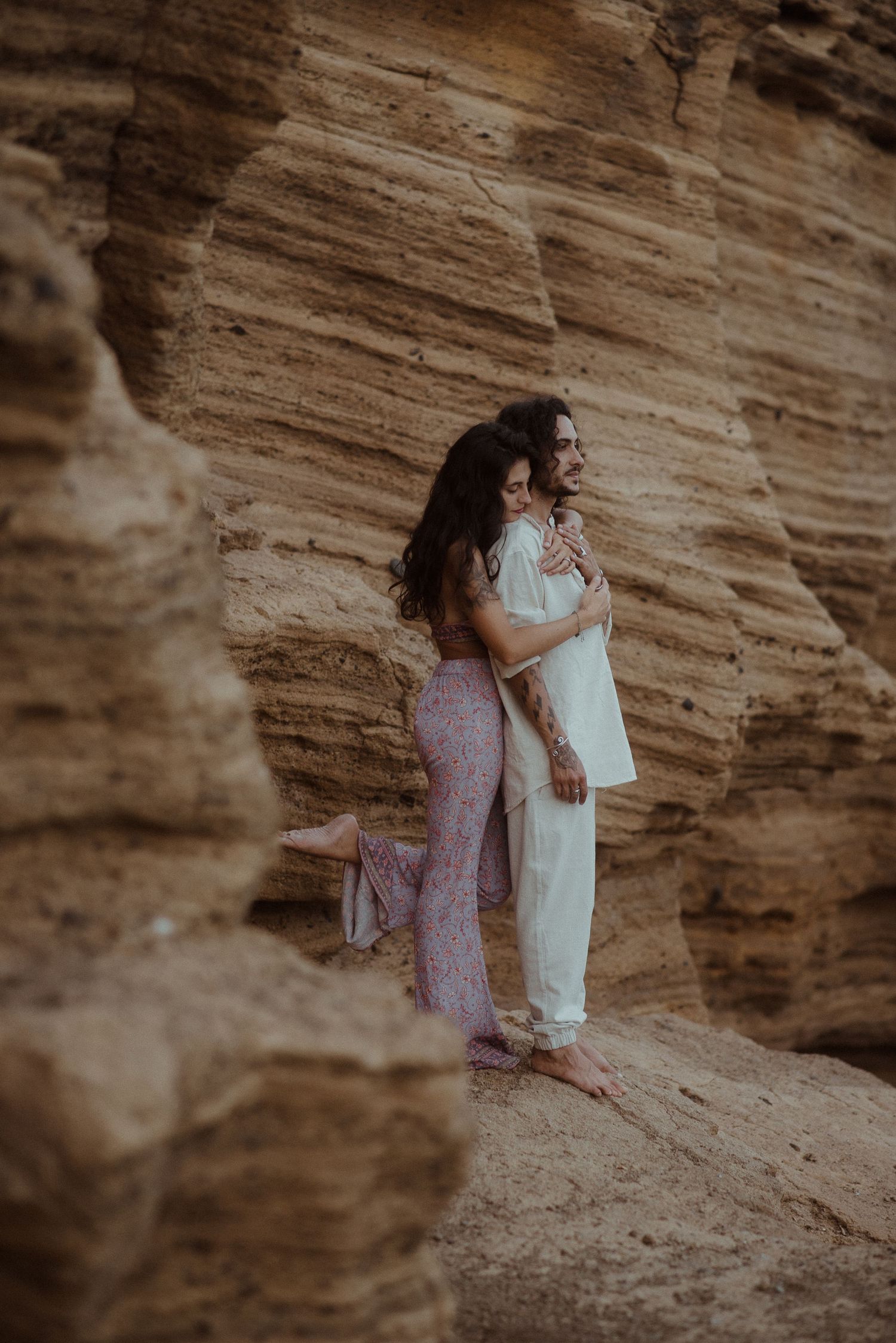 Two people share an intimate moment against layered sandstone cliffs by the sea.