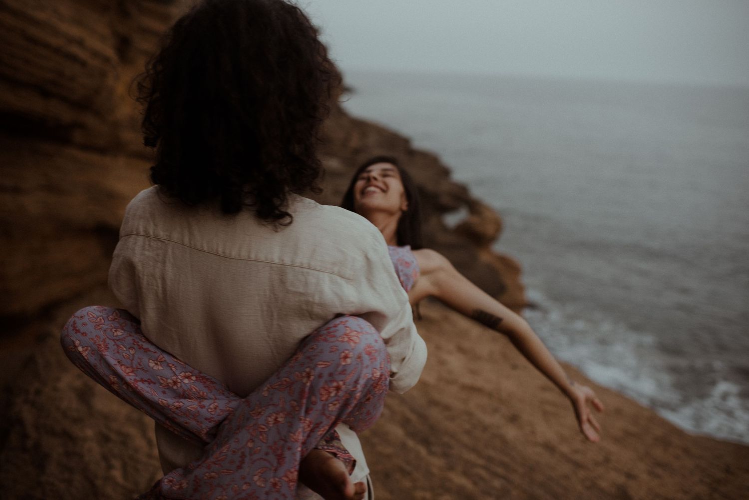 Couple playfully poses on rugged oceanside cliff during moody photoshoot.