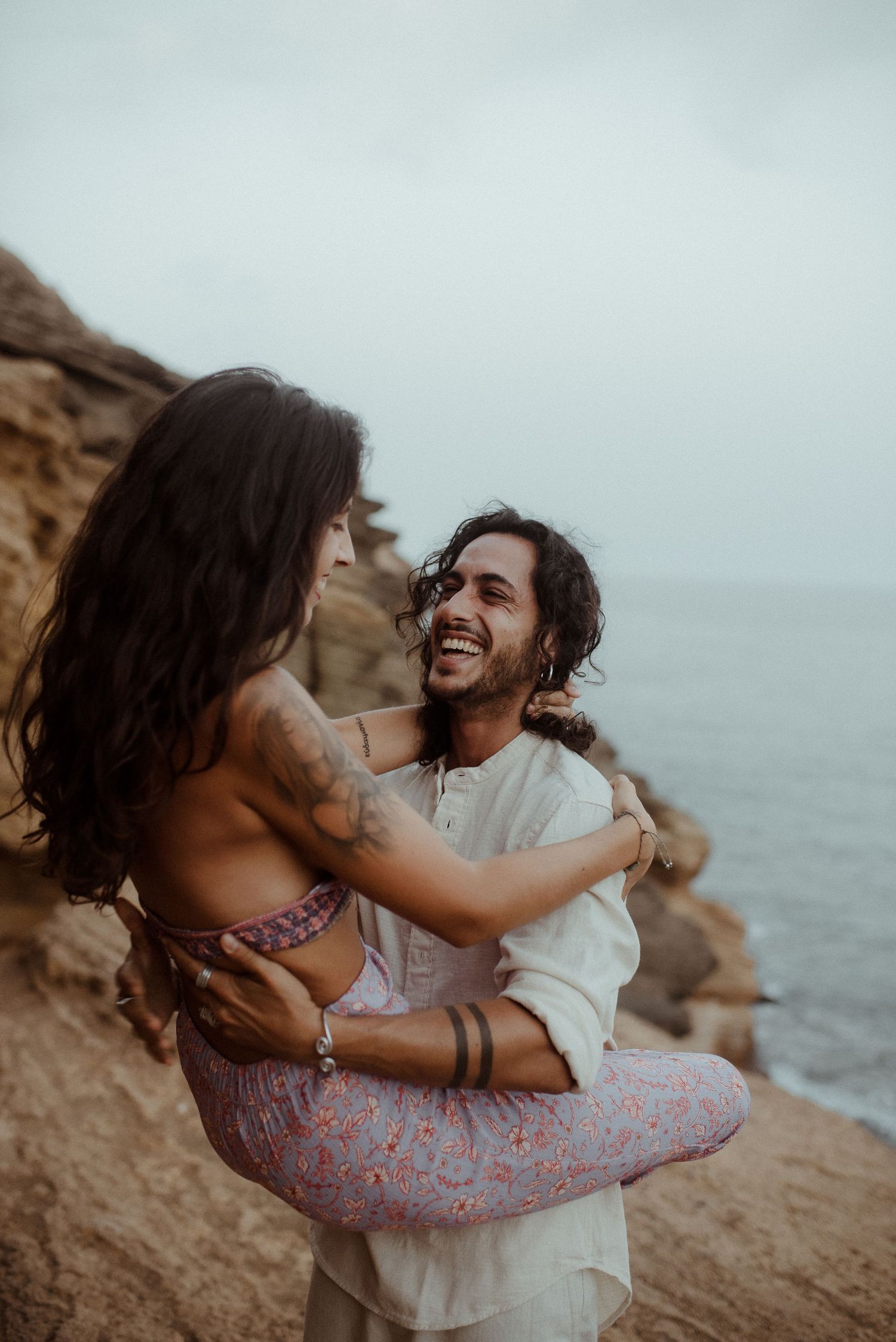 Joyful couple laughing while embracing on coastal cliff edge during sunset.