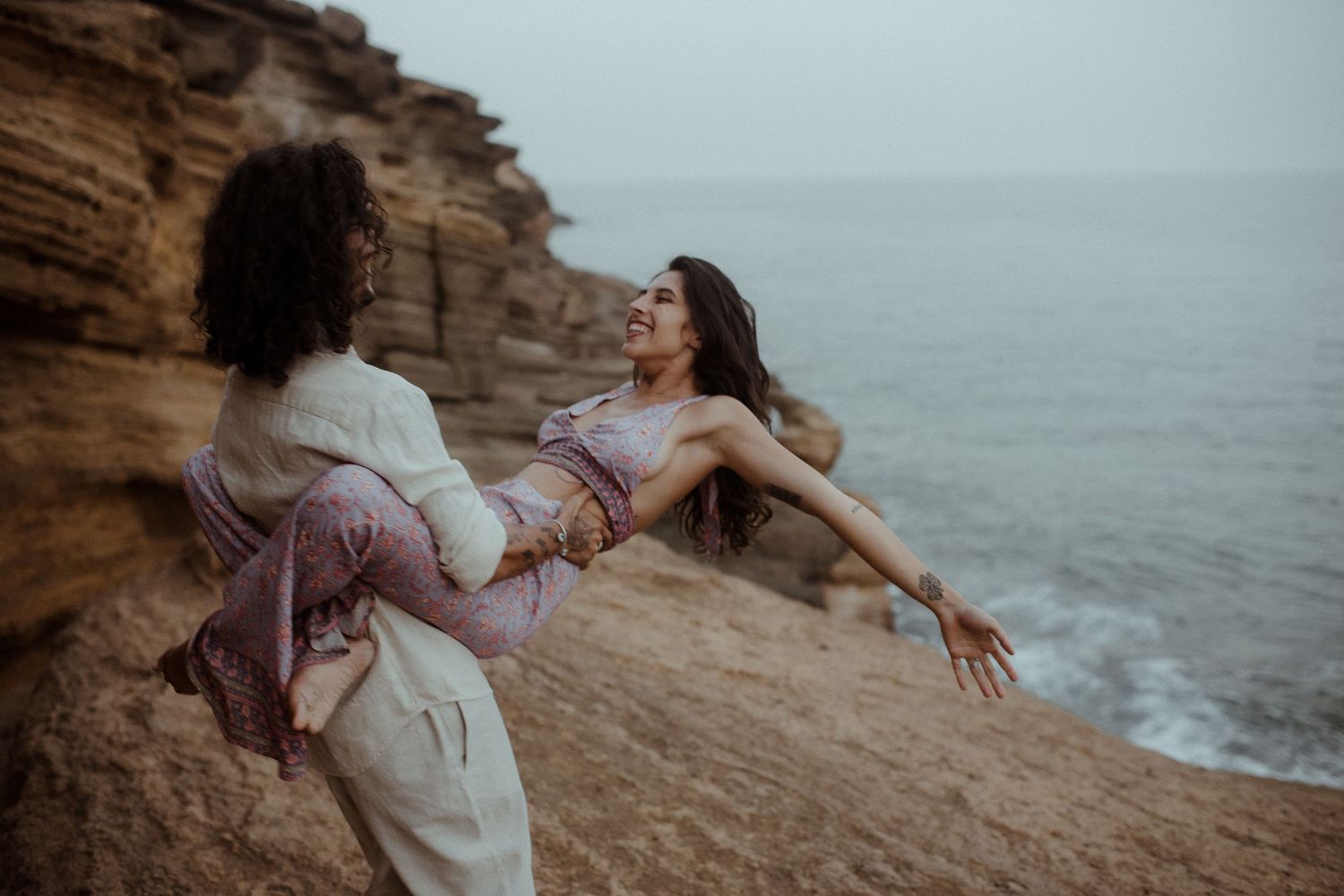 Two people strike dramatic dance pose on rocky ocean cliff at dusk.