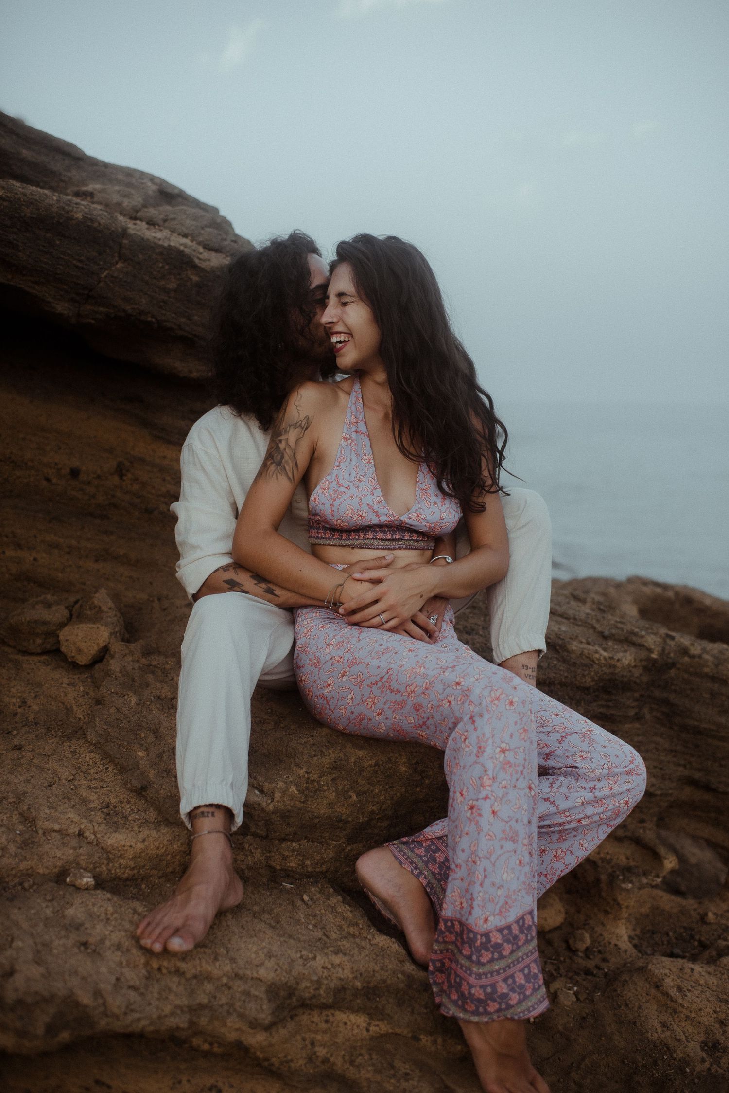 Intimate couple embracing on rocky cliff during sunset in matching light-colored outfits.