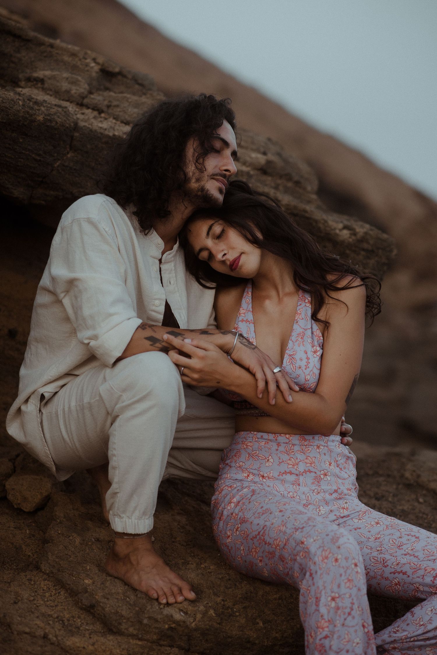Tender moment between couple resting on rocky cliff in casual bohemian attire.