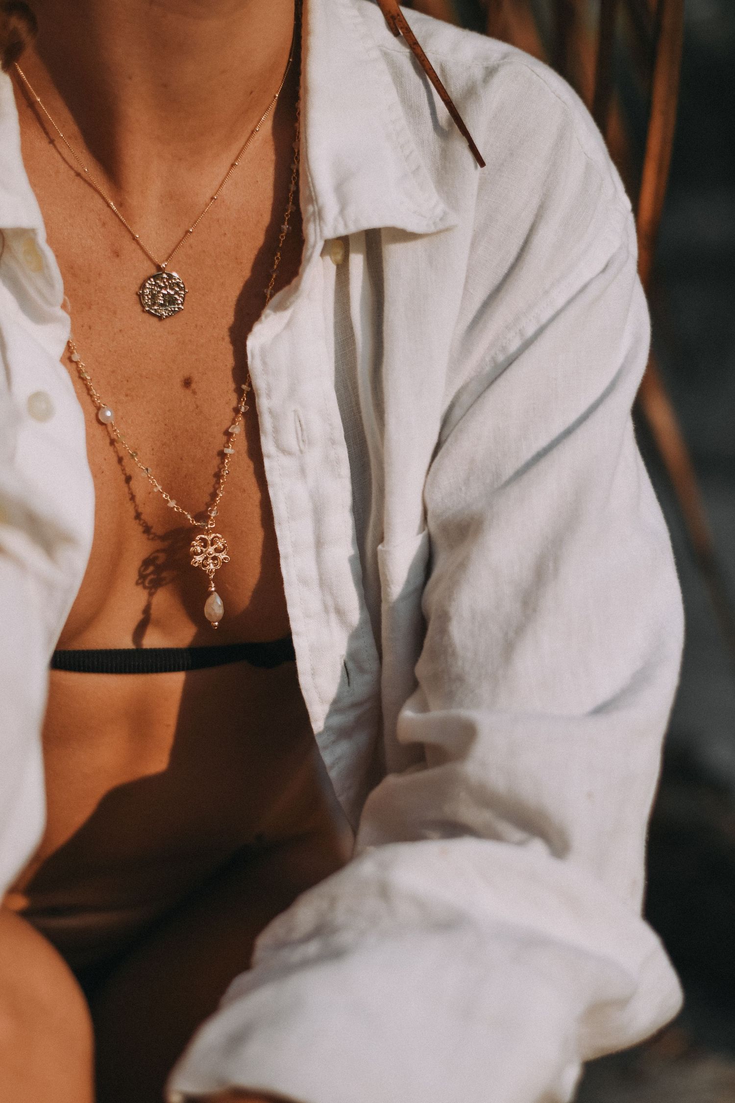 Close-up view of delicate layered necklaces with ornate pendants worn with a white button-down shirt and black undergarment.