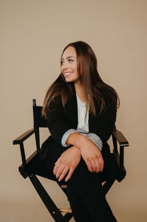 Professional headshot in director's chair wearing black blazer against neutral backdrop.