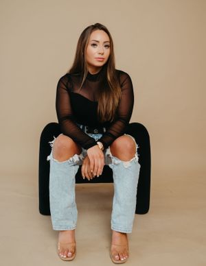 Fashion portrait of someone wearing a black mesh top and distressed jeans while sitting on a dark chair.