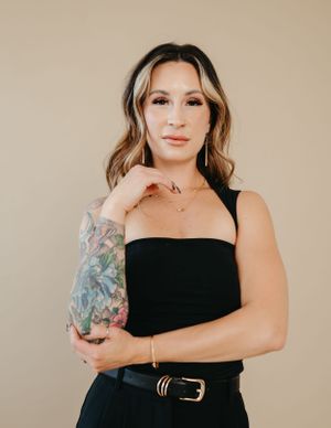 Studio portrait against beige backdrop showing fashionable black top and colorful arm tattoo.