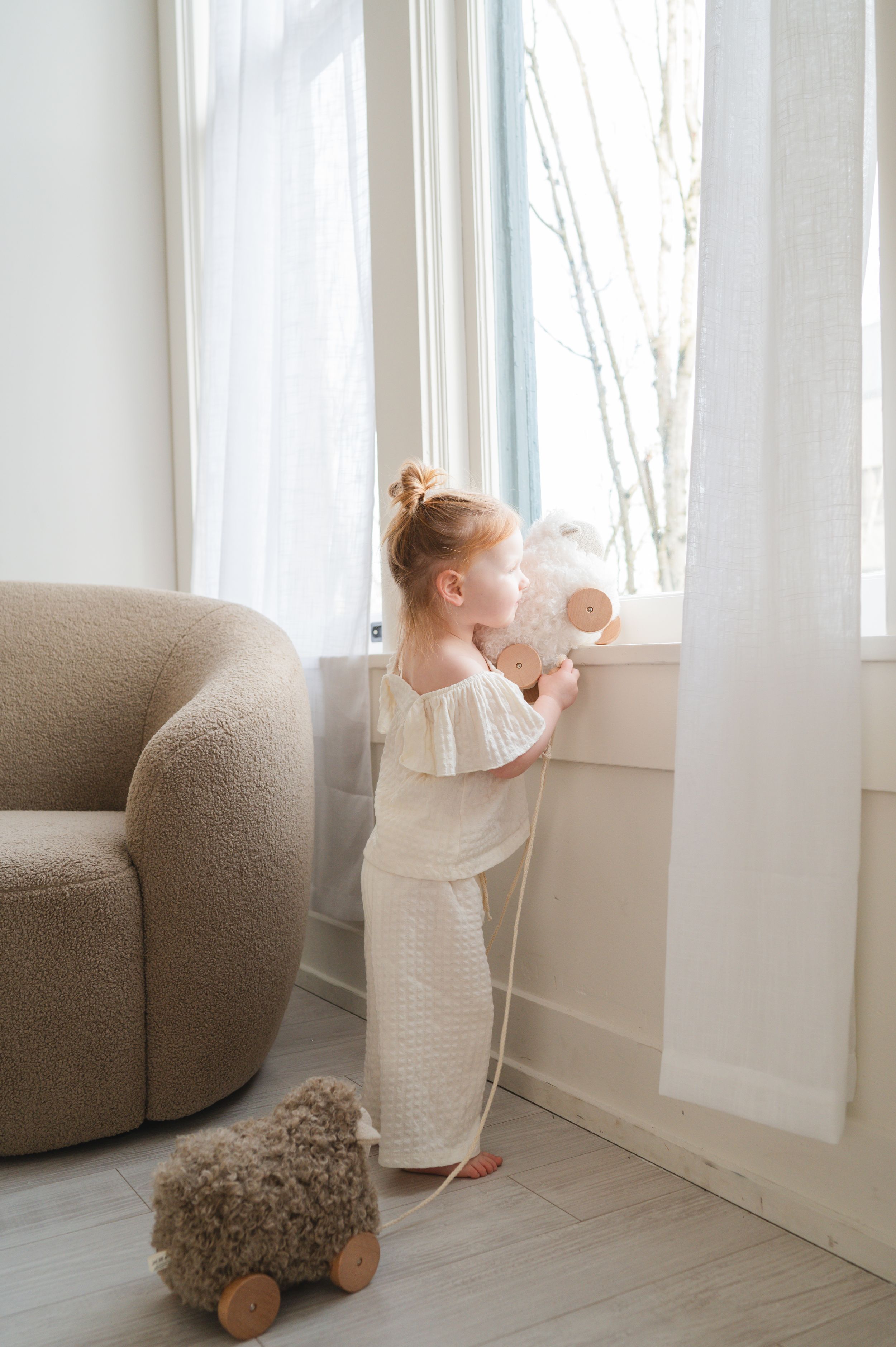 A toddler in a cream dress looks out a bright window next to a plush sheep toy in a minimalist room.