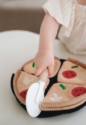 A small hand reaches for a plush pizza toy made from soft felt with pepperoni and cheese details.