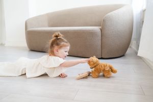 Young child in white dress plays with orange plush toy cat on light wooden floor beside curved beige sofa.