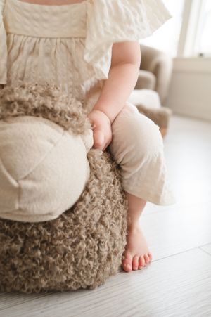 Small bare feet and white dress resting on fluffy brown floor cushion in bright sunlit room.