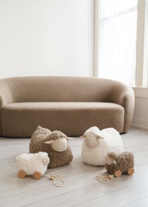 Plush sheep toys in brown and white colors arranged on a wooden floor next to a beige curved sofa near a window.