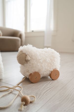 A fuzzy white wooden pull-along sheep toy with wheels sits on a light wood floor near a beige armchair.