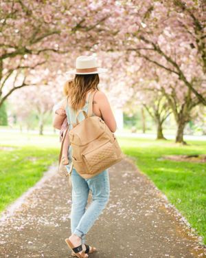 Person walking down path lined with blooming cherry blossom trees wearing casual attire and backpack.
