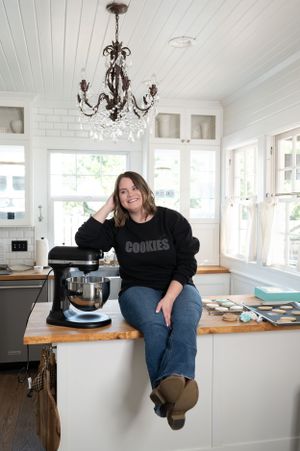 A bright white kitchen with a chandelier features someone sitting on a butcher block counter near a stand mixer.