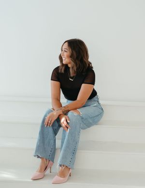 A woman in a black shirt and jeans sits casually on a white chair.