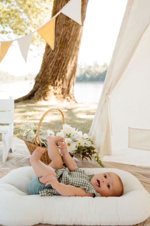 A baby lying on a white cushion near a tent with bunting and trees in background.