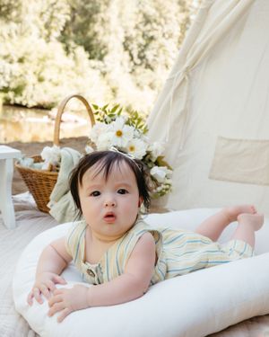 Baby relaxing on white bedding outdoors with picnic basket and flowers in the background.