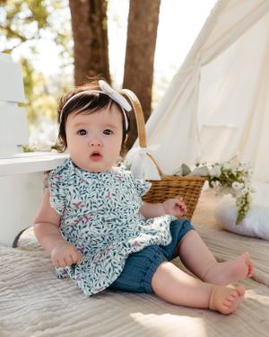 A baby wearing a floral dress and headband sits on a blanket near a white tent.