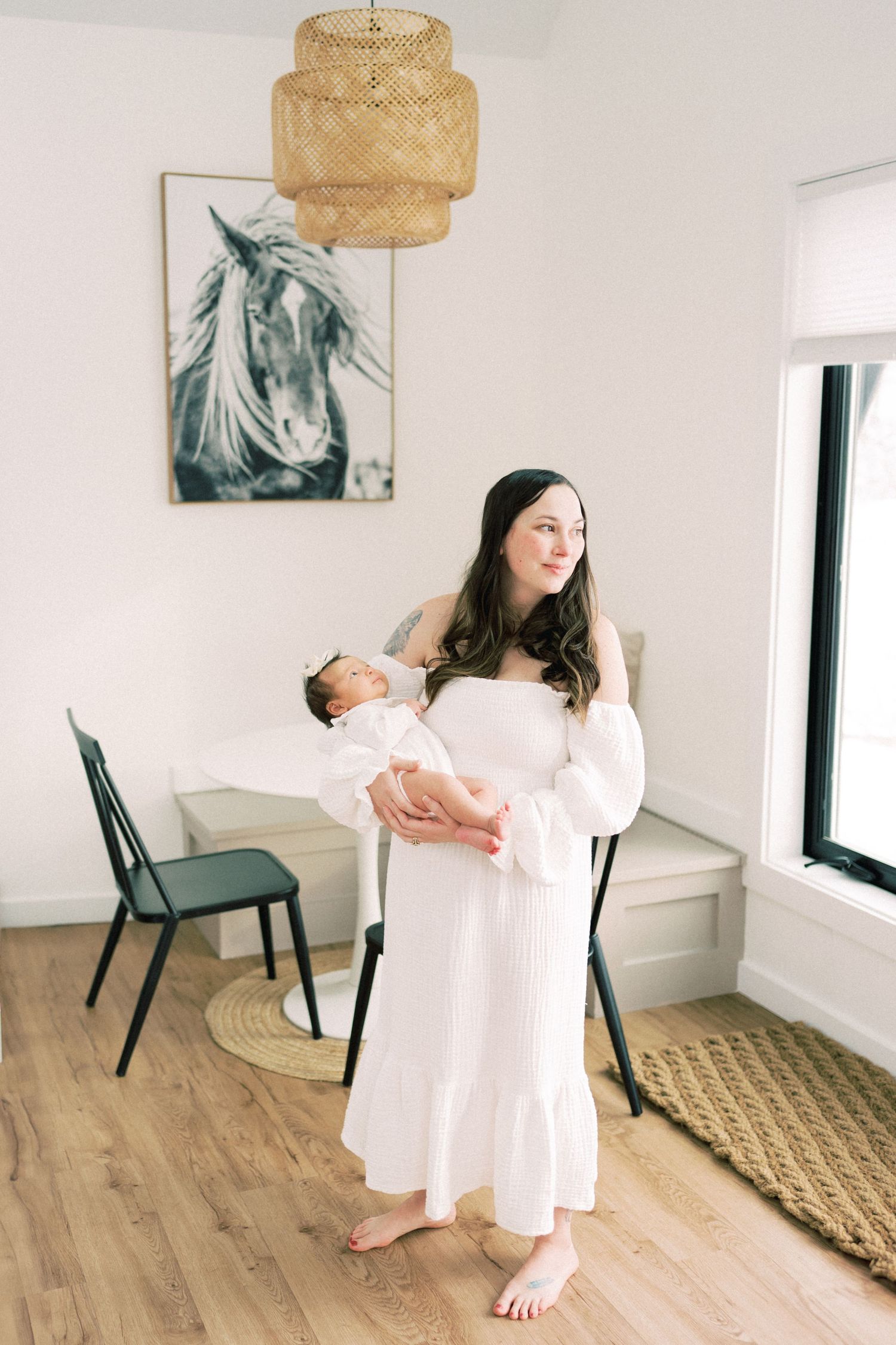 Parent holding baby in white dress near horse artwork and woven pendant light.