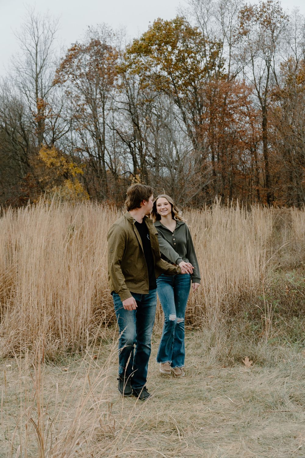 A couple walks through tall autumn grass with fall foliage in the background.