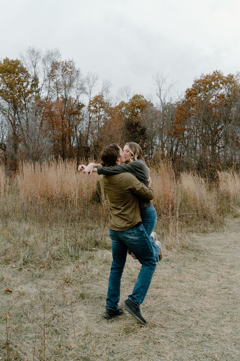 Couple embracing playfully in a fall field with dried tall grass and autumn trees in the background.