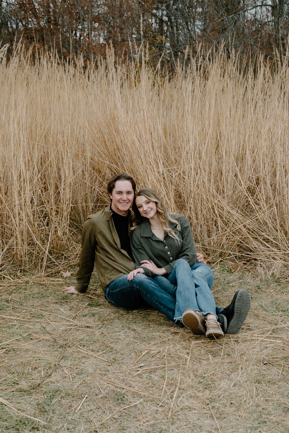 A couple sits together in tall autumn grass wearing casual clothing for an outdoor photoshoot.