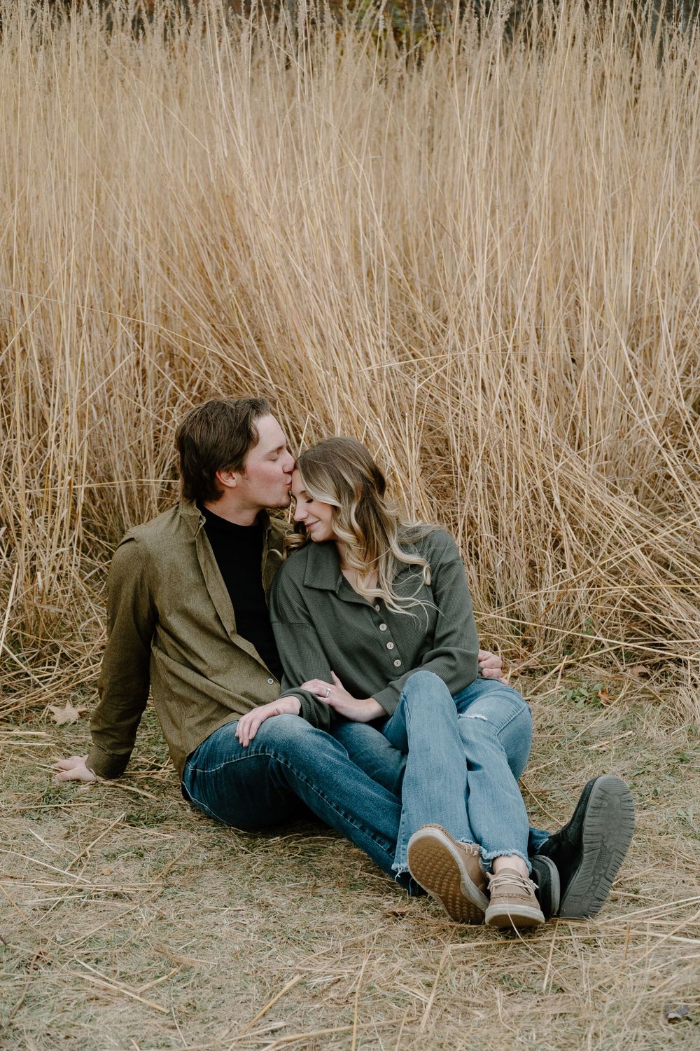 A romantic couple shares an intimate moment while sitting in tall dried grass during a photo session.