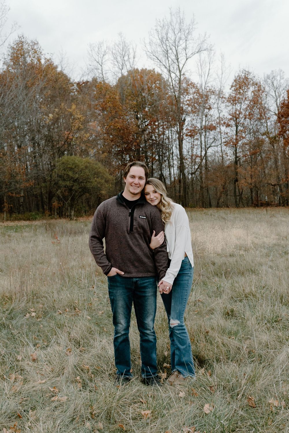 Couple standing together in a rustic field surrounded by fall foliage and bare trees.