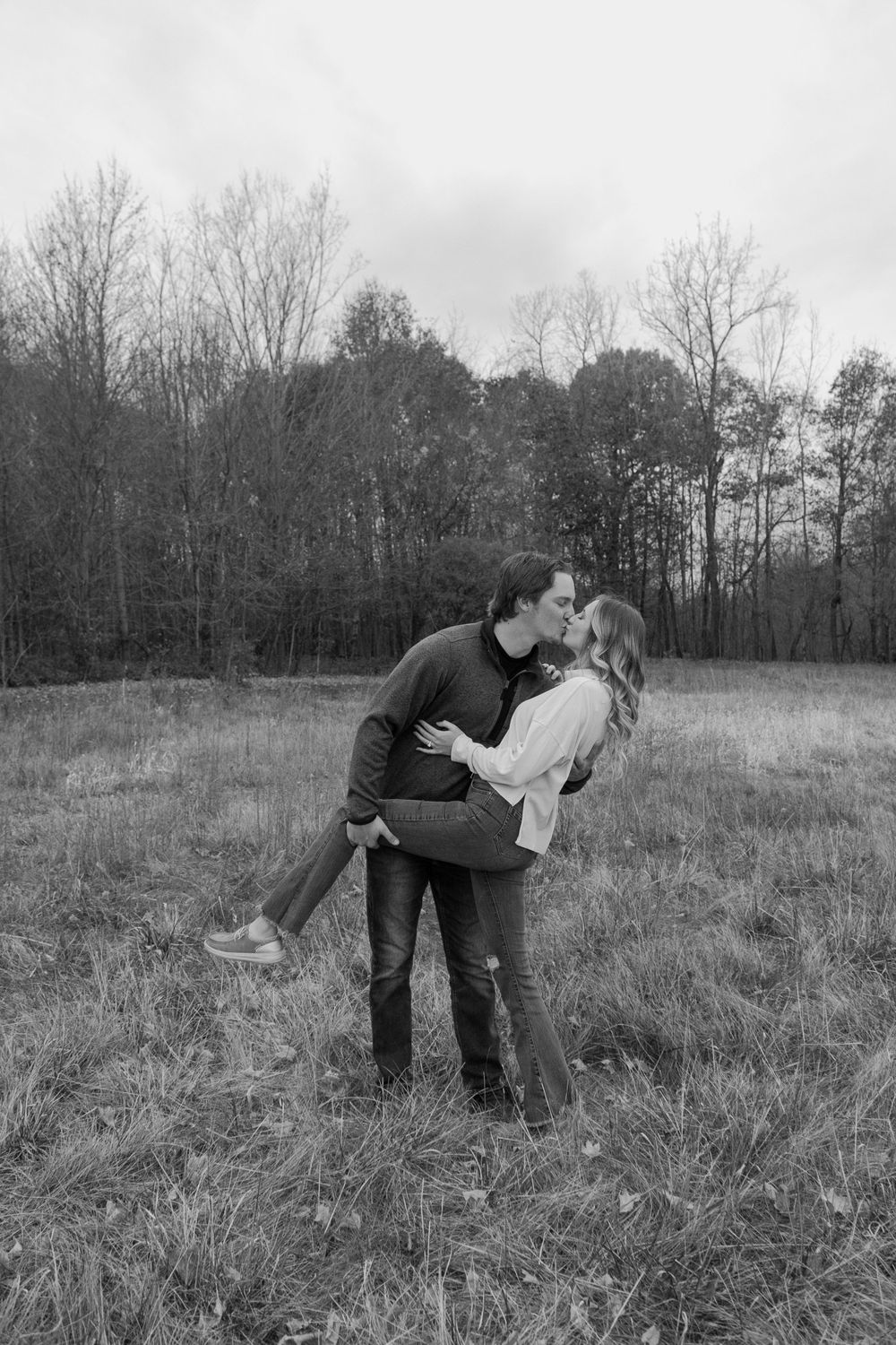 A romantic black and white photo of a couple embracing in a field with bare trees in background.