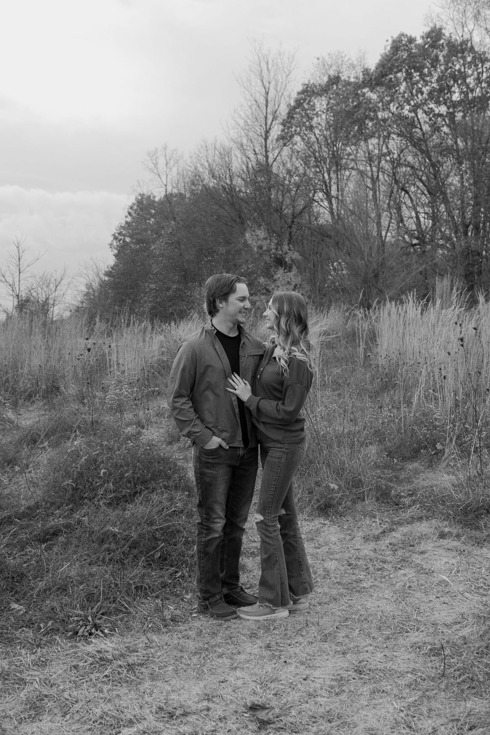 Couple shares tender moment standing in field with tall grass and winter trees behind them.