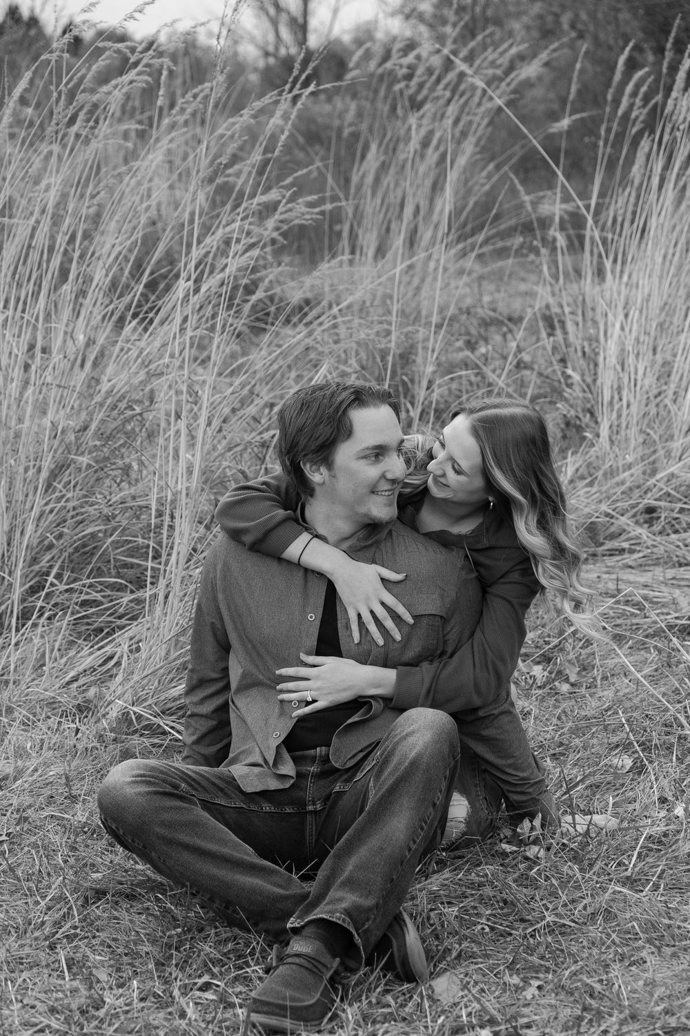 Black and white portrait of couple embracing in tall grass during outdoor photo session.