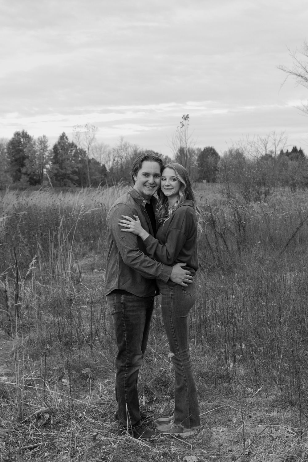 Black and white photo of couple embracing while standing in a winter field.