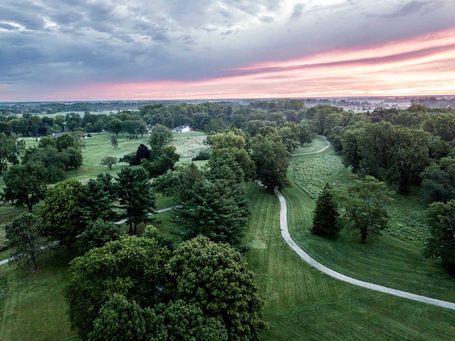 Aerial view of a golf course with winding cart paths and lush trees under a dramatic pink and purple sunset sky.