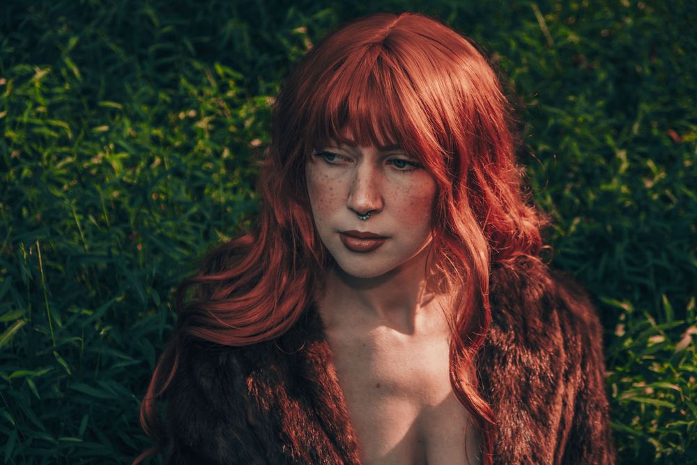 Close-up portrait with vibrant red hair against lush green natural background.