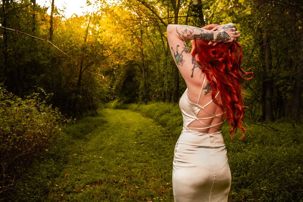 Figure in fitted dress with bright hair poses in sunlit forest path.