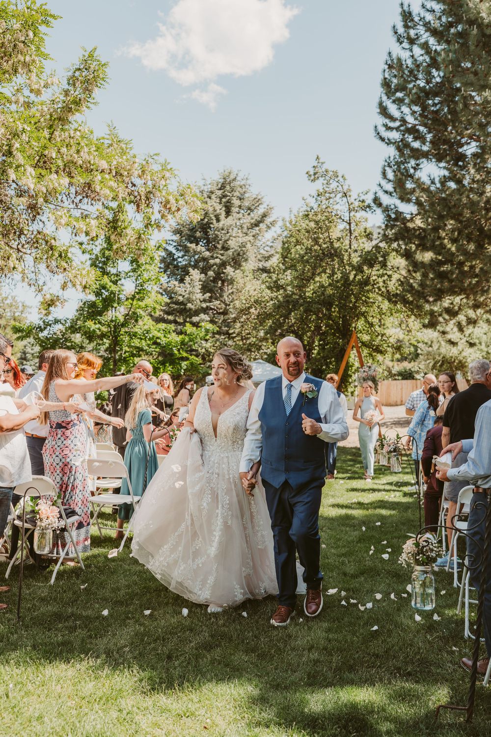 A wedding ceremony celebration in a sunlit garden with guests throwing flower petals as a couple walks down the aisle.
