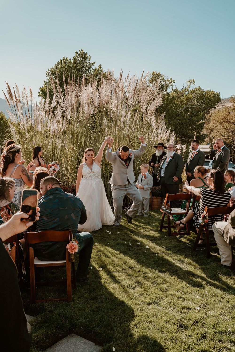 An outdoor wedding ceremony taking place in a garden setting with tall ornamental grasses and golden sunlight.