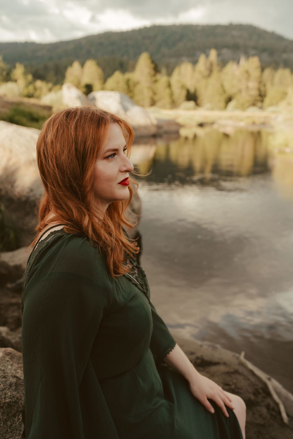 A redhead in a dark coat sits by a scenic lake with dramatic rock formations and moody cloudy skies.