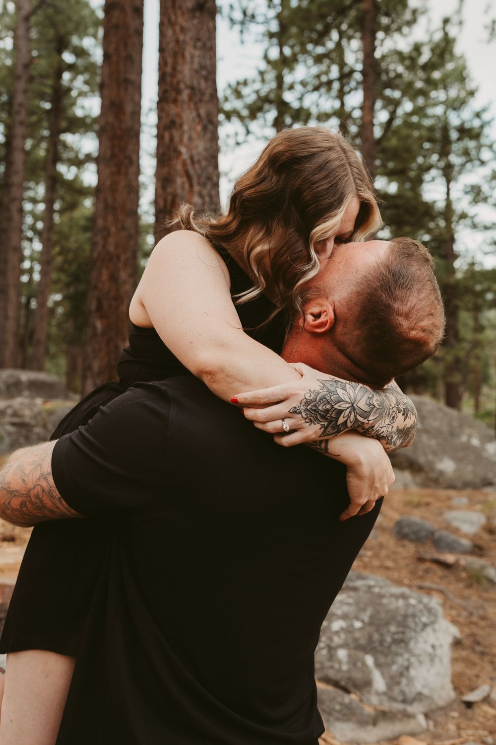 Couple sharing an intimate embrace in a forest setting with pine trees and rocky ground in warm sunset lighting.