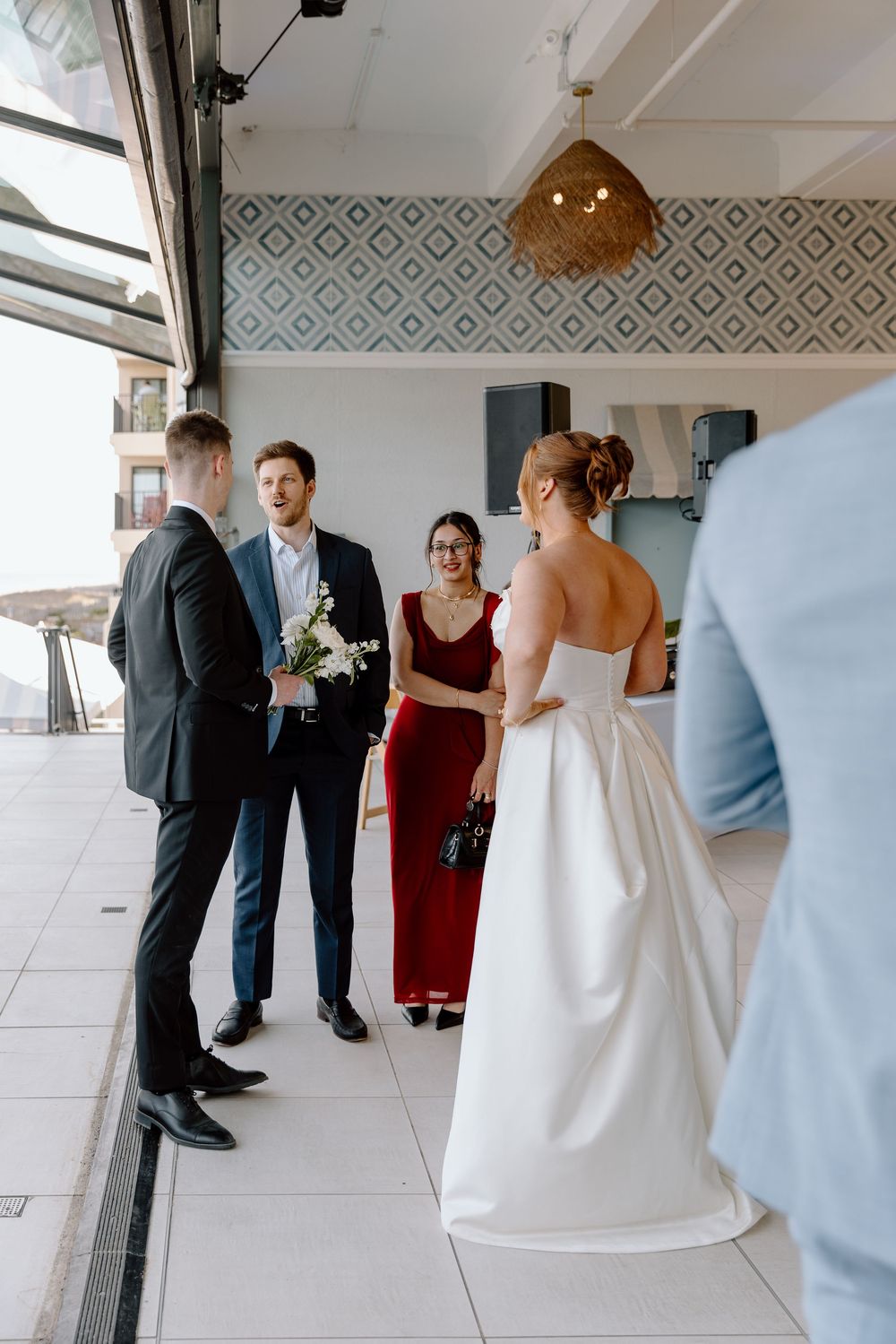 Wedding guests gather on a balcony with geometric wallpaper and modern pendant lighting during a celebration.