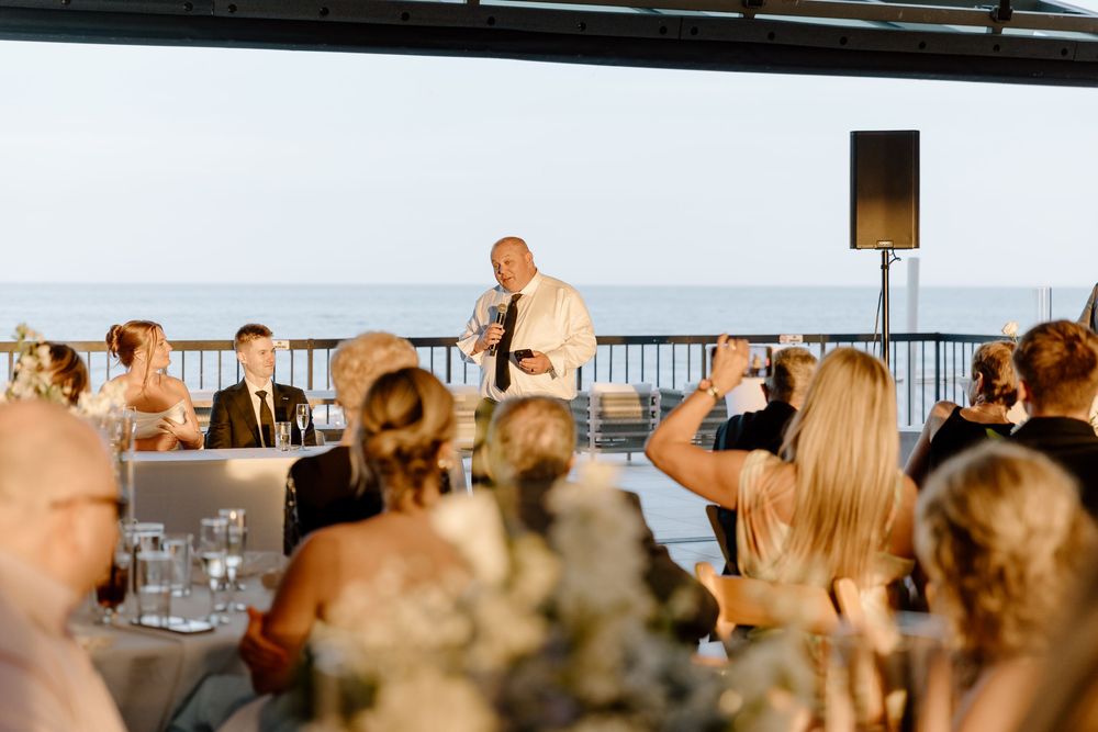 A speaker addresses guests during an outdoor reception on a deck overlooking water at sunset.