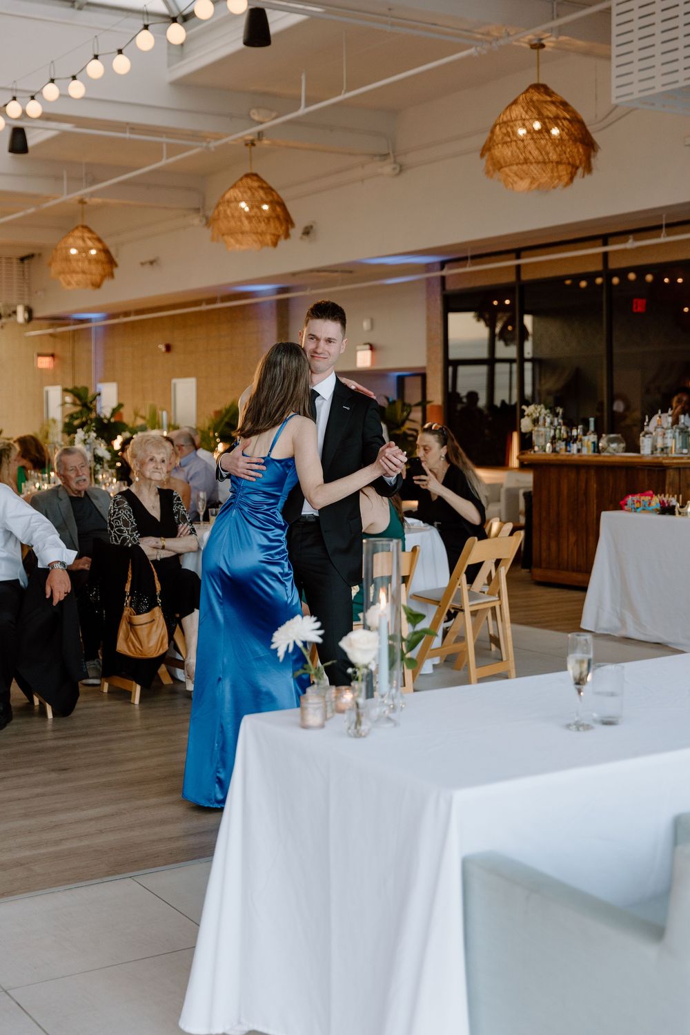 A banquet hall set with white tablecloths features elegant gold pendant lights and wedding guests enjoying drinks.